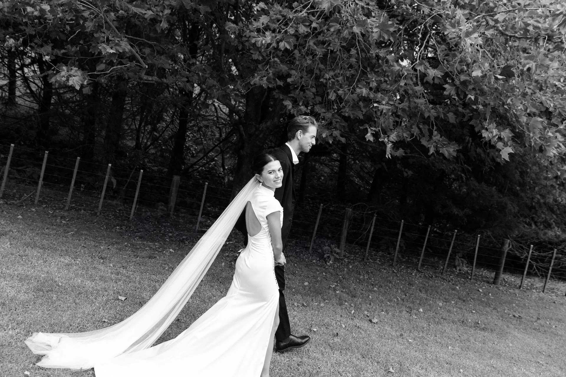A black and white photo of a bride and groom walking outdoors, with the bride in a white wedding dress and veil, and the groom in a black suit, smiling and holding hands.
