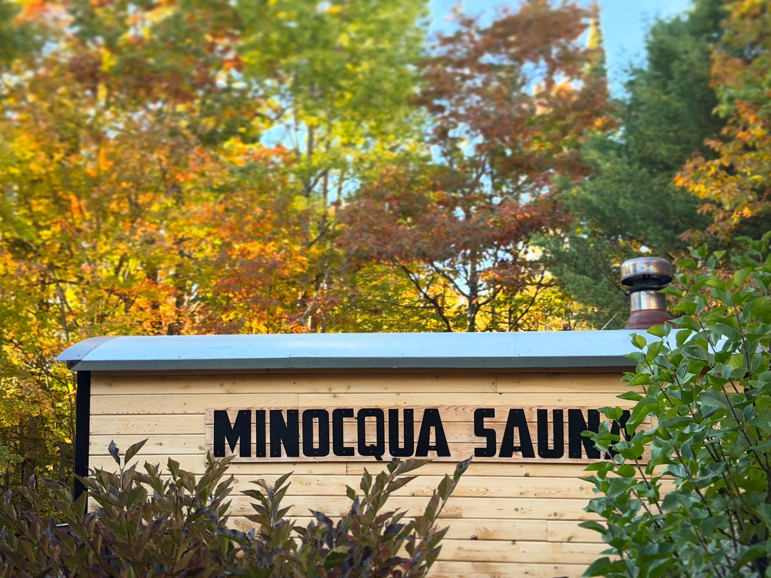 Minocqua Sauna mobile trailer sauna unit, traditional wood burning sauna on a trailer parked amongst a beautiful fall sunny day with leaves changing a multitude of different colors in the foreground and background, framing the cedar wood sauna