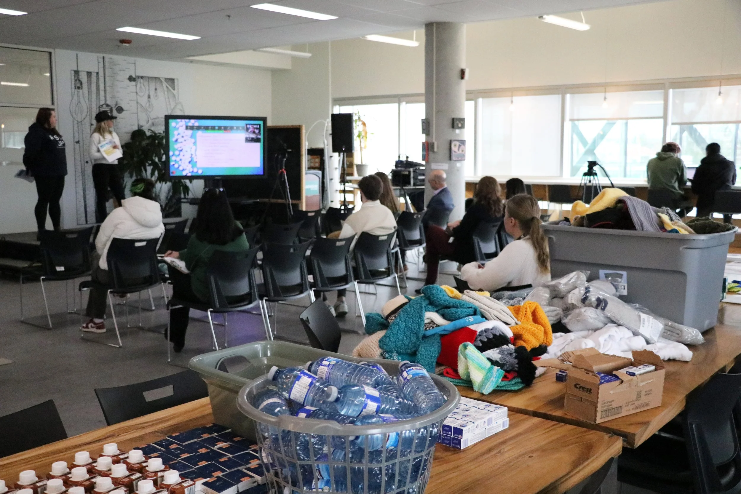 People seated in a meeting or presentation room with some sitting and others standing at the front near a large screen, with tables filled with various items like water bottles, boxes, and clothing.