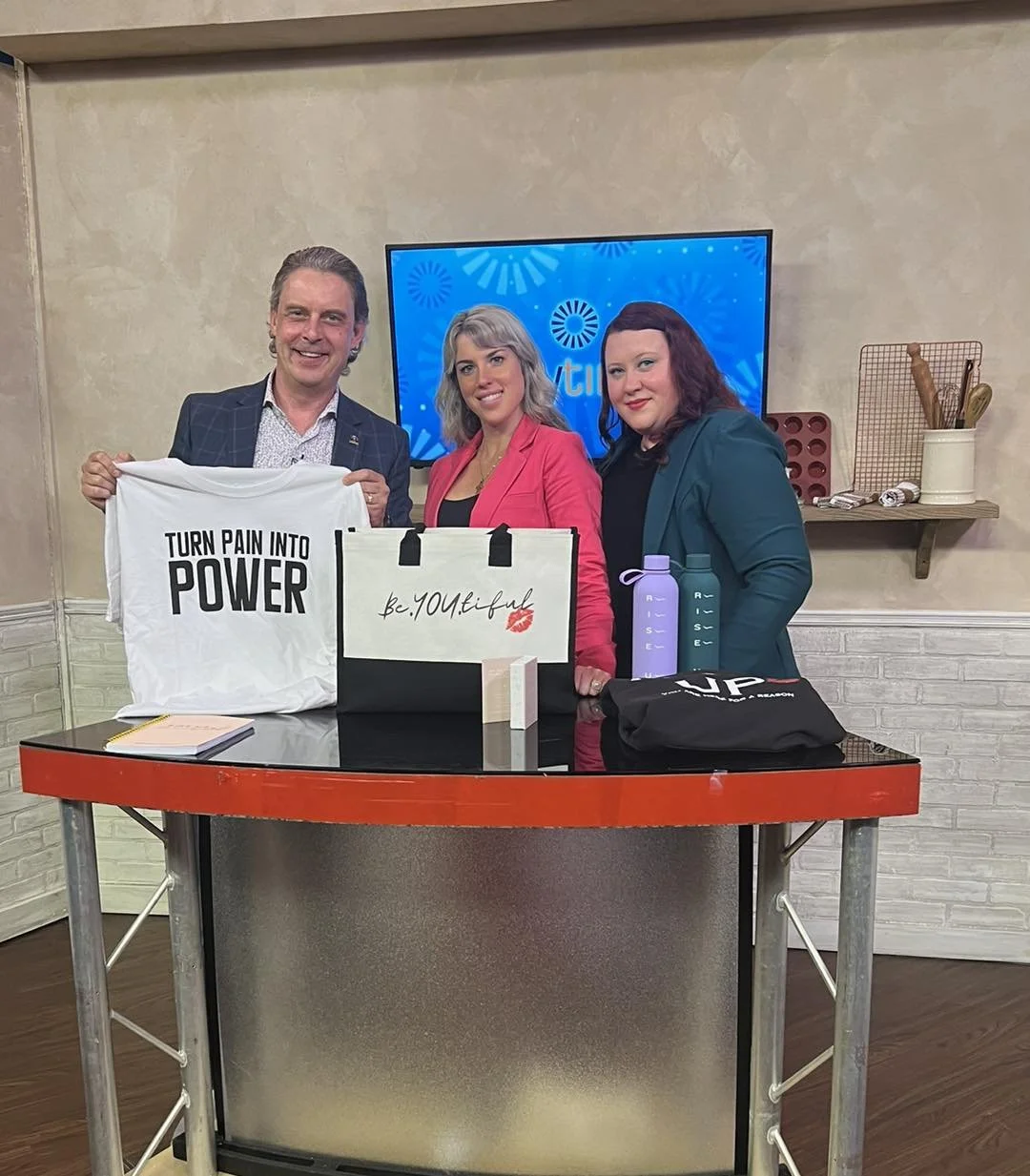 Three people standing behind a table displaying gifts and promotional items, with a television screen in the background. The man on the left is holding a white t-shirt that reads "TURN PAIN INTO POWER." The woman in the middle is wearing a pink blazer, and the woman on the right is wearing a teal blazer. There are water bottles, a tote bag with the text "be.YOU.tiful," and other items on the table.”}】