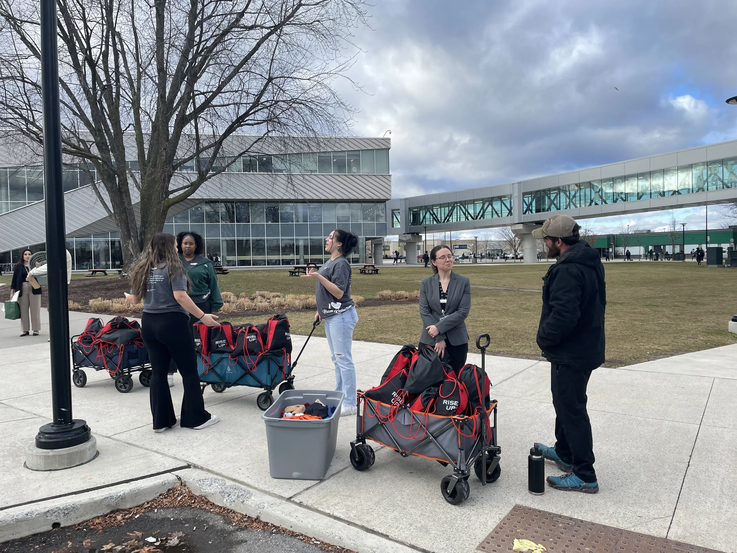 Group of people standing outdoors near a modern building with glass walls and a skybridge, some with shopping and activity carts.