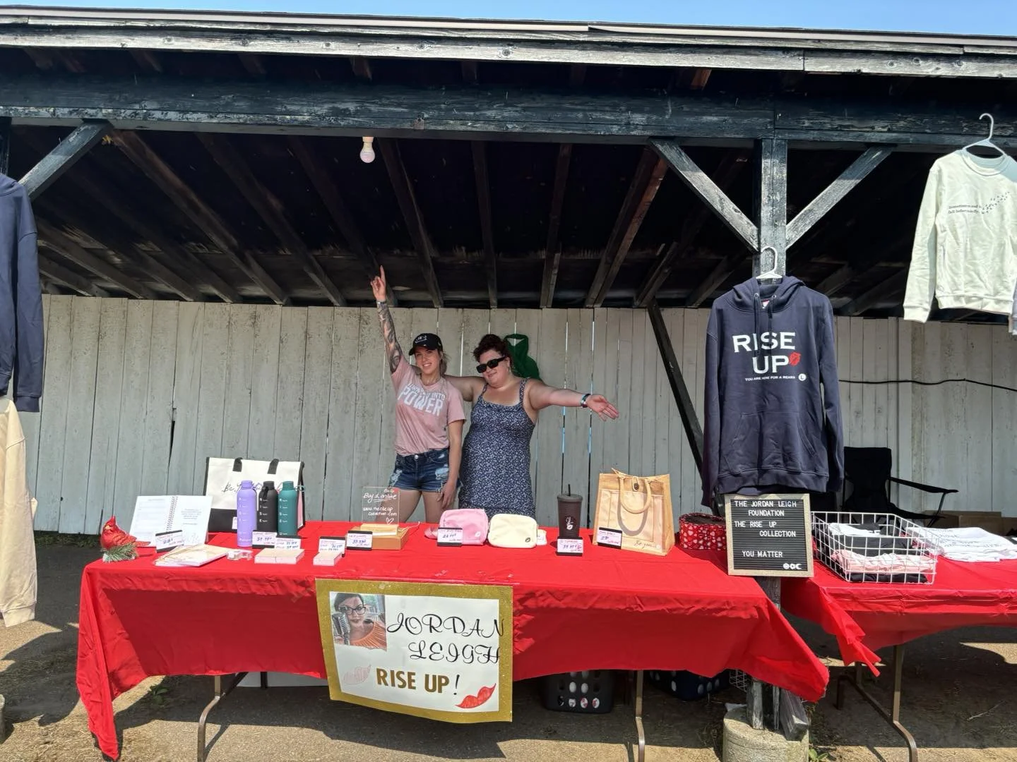 Two women standing behind a table at a fundraising booth under a wooden pavilion. The table is covered with a red tablecloth and displays various items for sale, including water bottles, a pink bag, and a sign that reads "Jordan Lugg RISE UP!" A hoodie with "RISE UP" printed on it hangs on the right side of the pavilion. The women are smiling; one is raising her arm and pointing toward the sky.