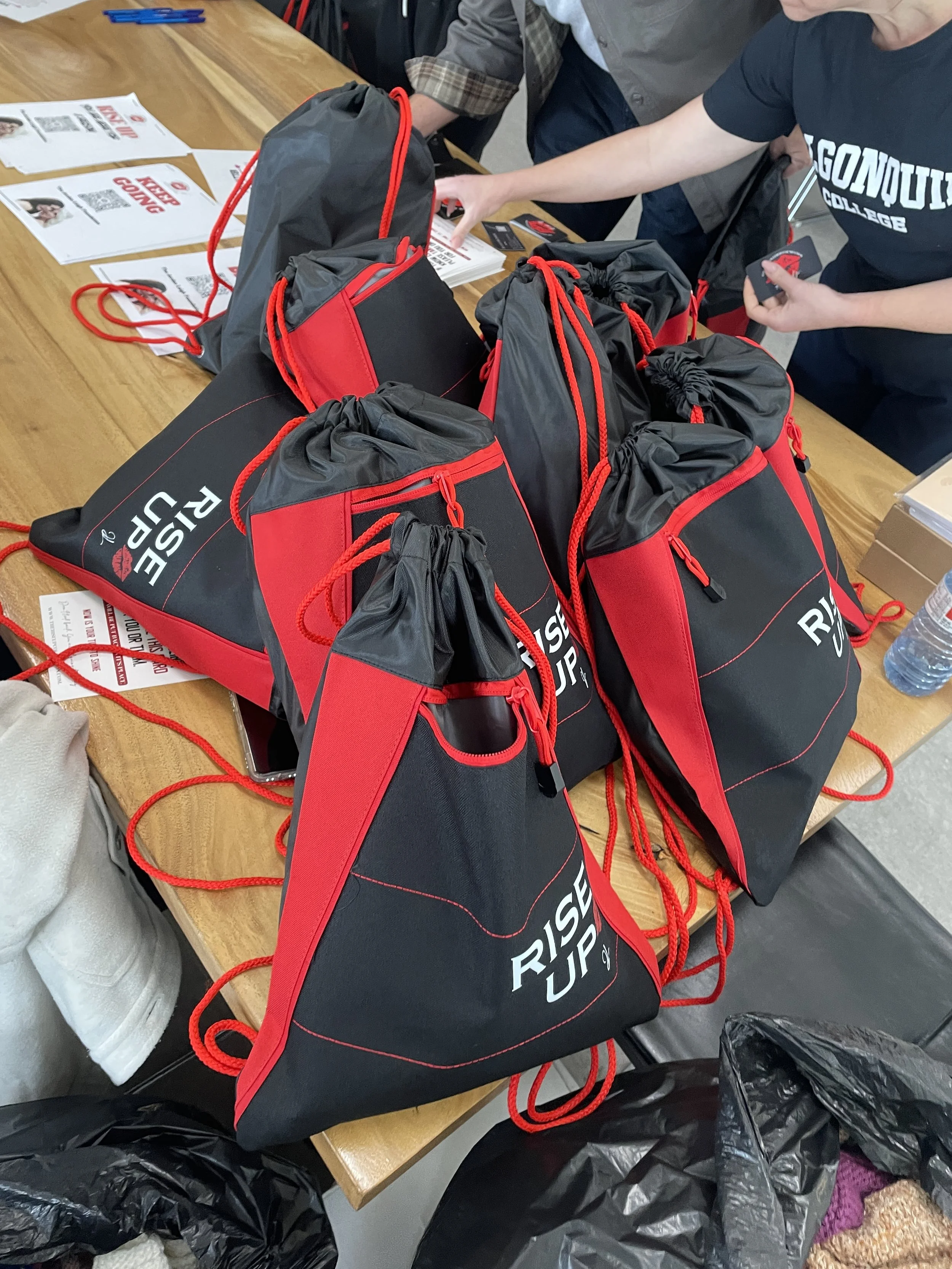 Multiple black and red drawstring bags on a wooden table with newspapers and water bottles nearby, with people reaching for the bags, and a person wearing a black T-shirt with white text.