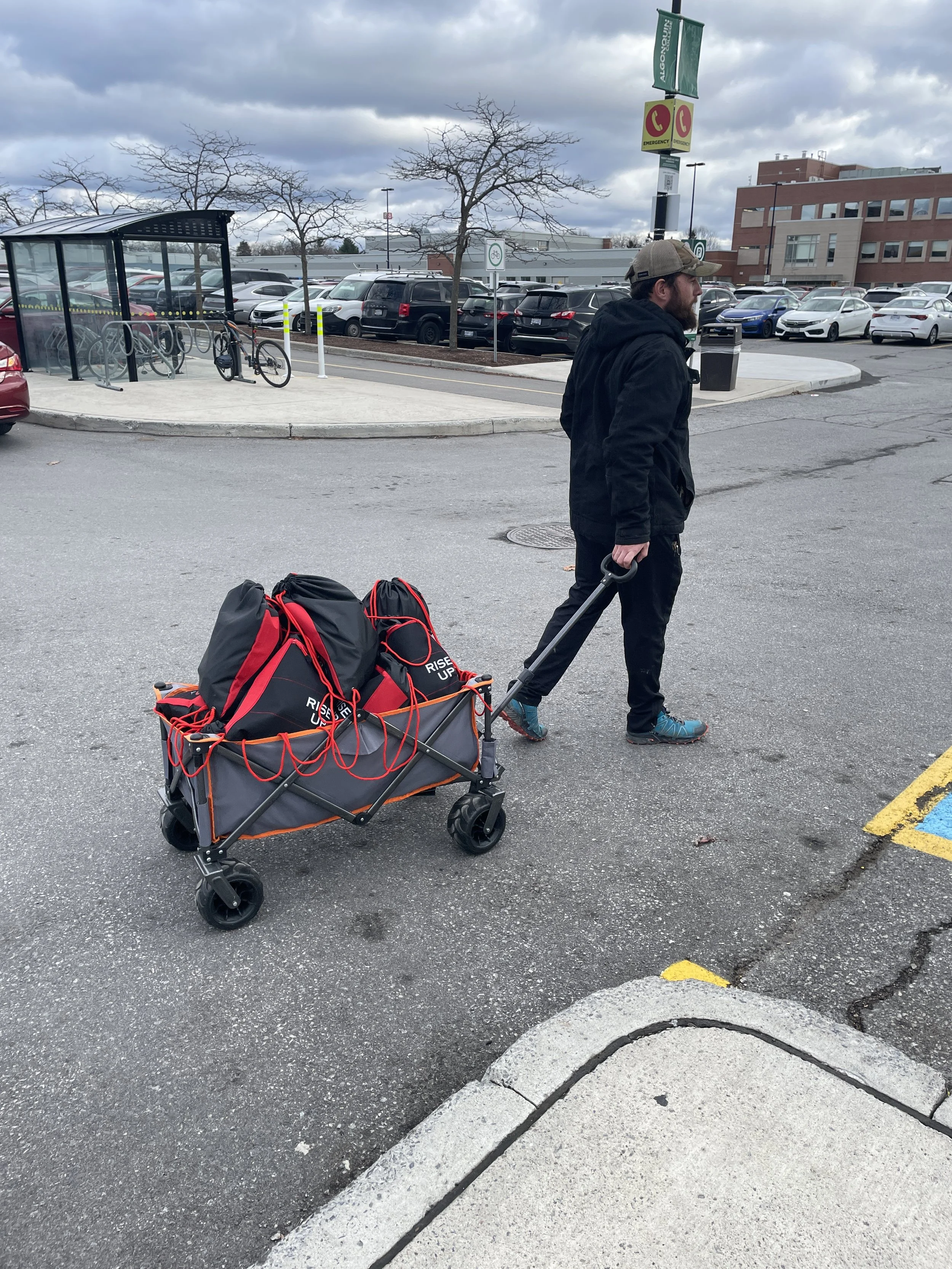 A man dressed in black with a cap walks across a parking lot pulling a collapsible wagon loaded with black and red backpacks.