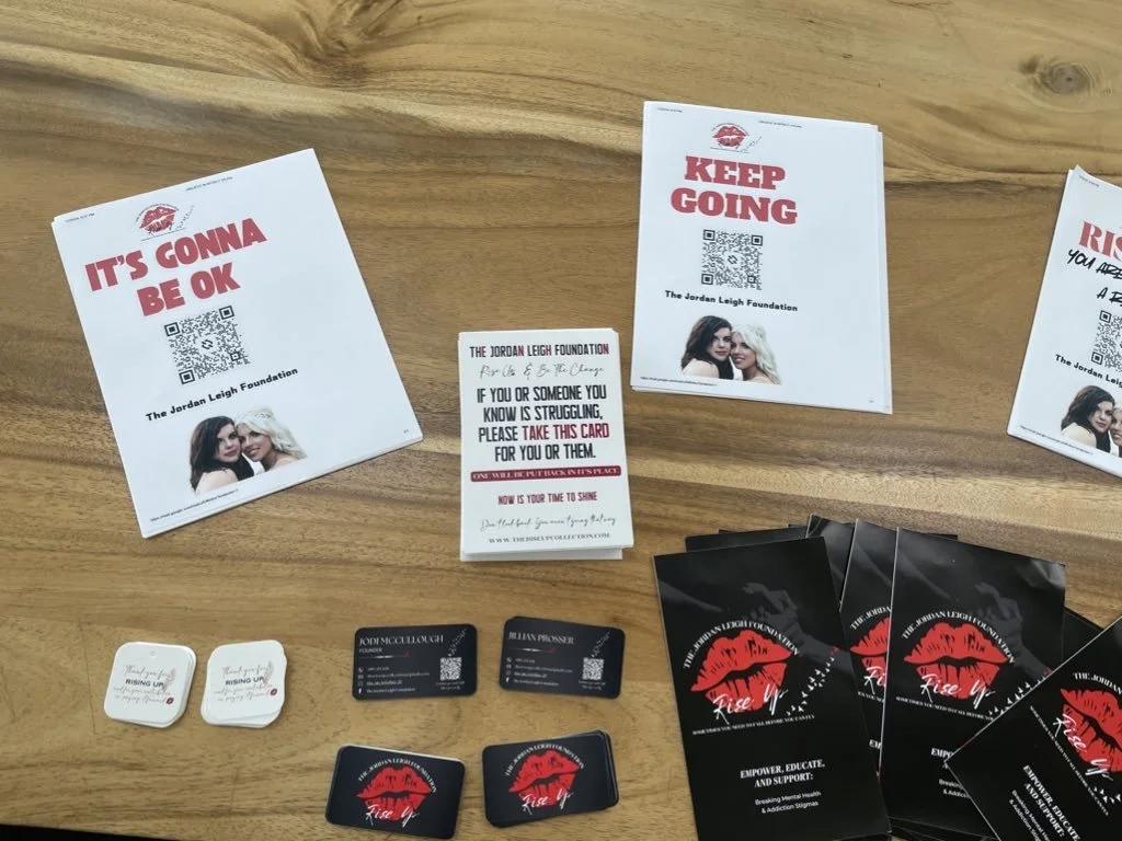 Flyers and cards related to The Jordan Leigh Foundation, with messages like 'It's Gonna Be OK' and 'Keep Going,' along with business cards and branded stickers, arranged on a wooden surface.