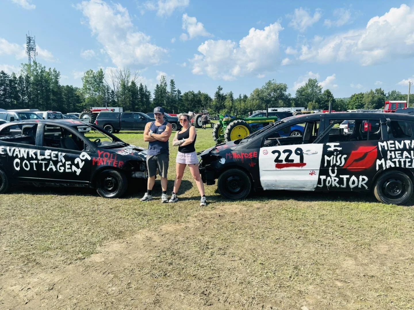 Two people standing between two painted cars with messages about mental health, parked on grass at an outdoor event with vehicles and tractors in the background.