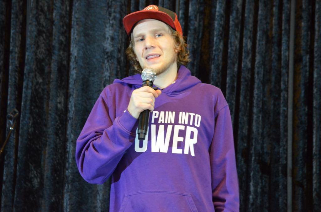 Young man in a purple hoodie with the phrase "Pain Into Power" on it, holding a microphone, standing in front of a dark curtain.
