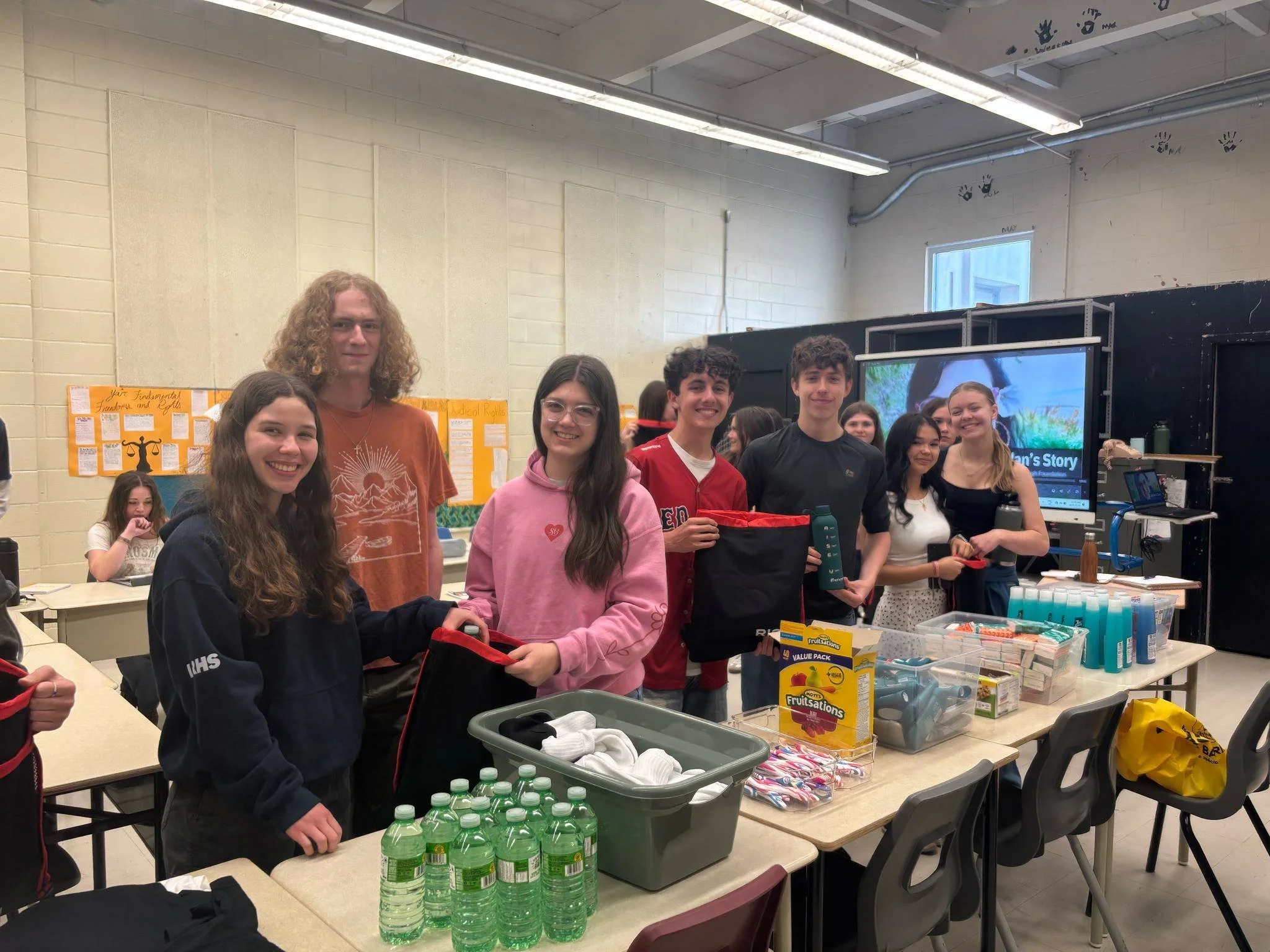 Group of seven teenagers standing around tables with supplies in a classroom or community center, smiling at the camera.