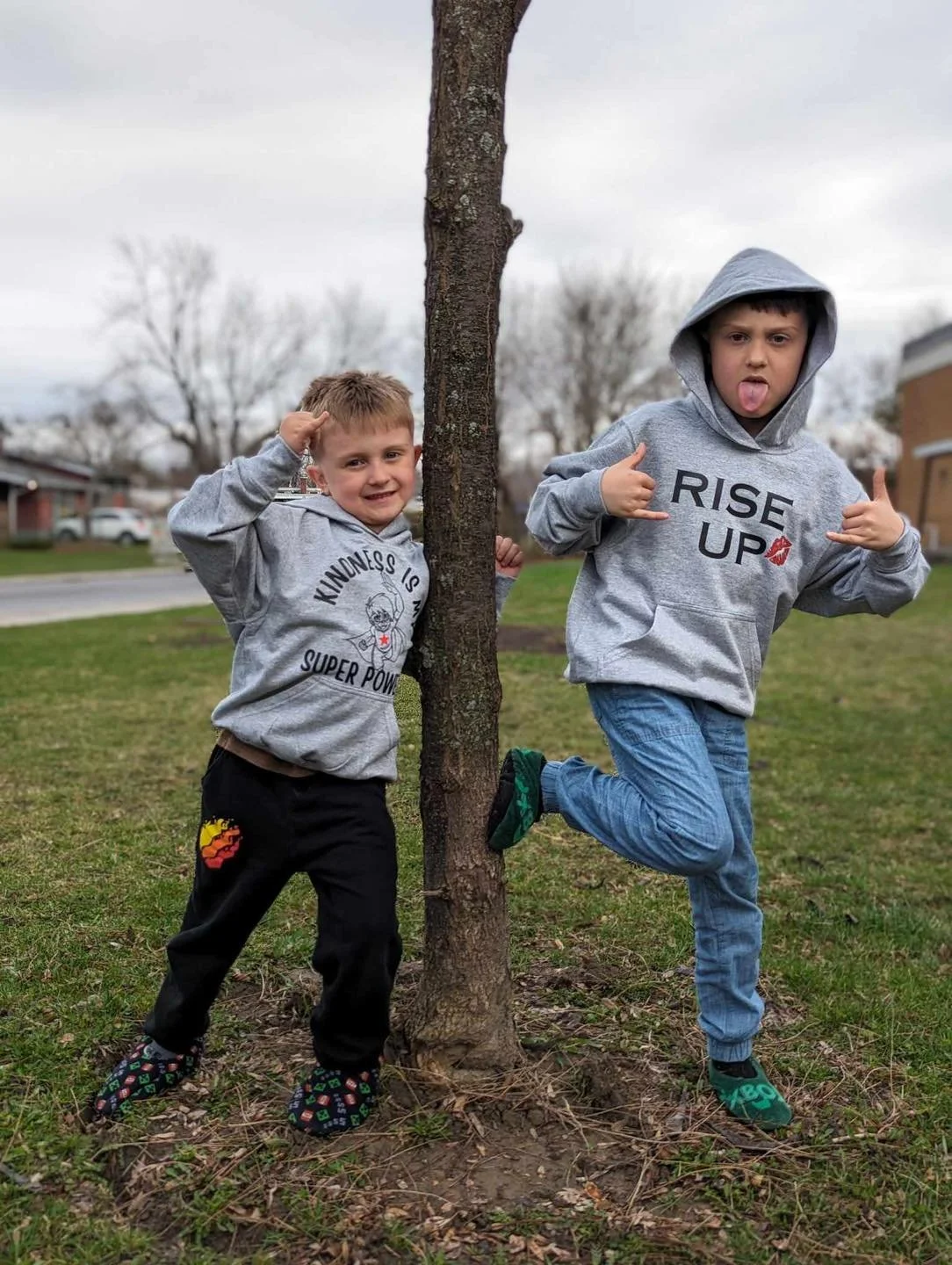 Two young boys posing playfully next to a small tree in a park. The boy on the left is smiling, wearing a gray hoodie and black pants with a colorful patch on the thigh. The boy on the right is making a silly face, sticking out his tongue, and wearing a gray hoodie with 'RISE UP' printed on it, blue jeans, and green sneakers. Both are holding up their hands with fingers making playful gestures.