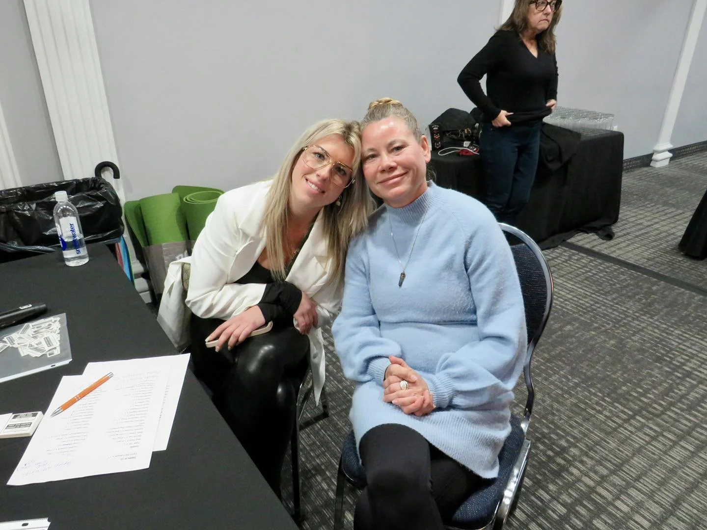 Two women sitting at a table, smiling and posing for the photo. One woman has blonde hair, glasses, and is wearing a white blazer, leaning her head toward the other woman. The other woman has her hair in a bun and is wearing a light blue sweater, sitting with her hands clasped. There are papers, a pen, and a water bottle on the table in front of them. In the background, there is a woman standing with her hands on her hips, and behind her are stacked water glasses and some black bags.