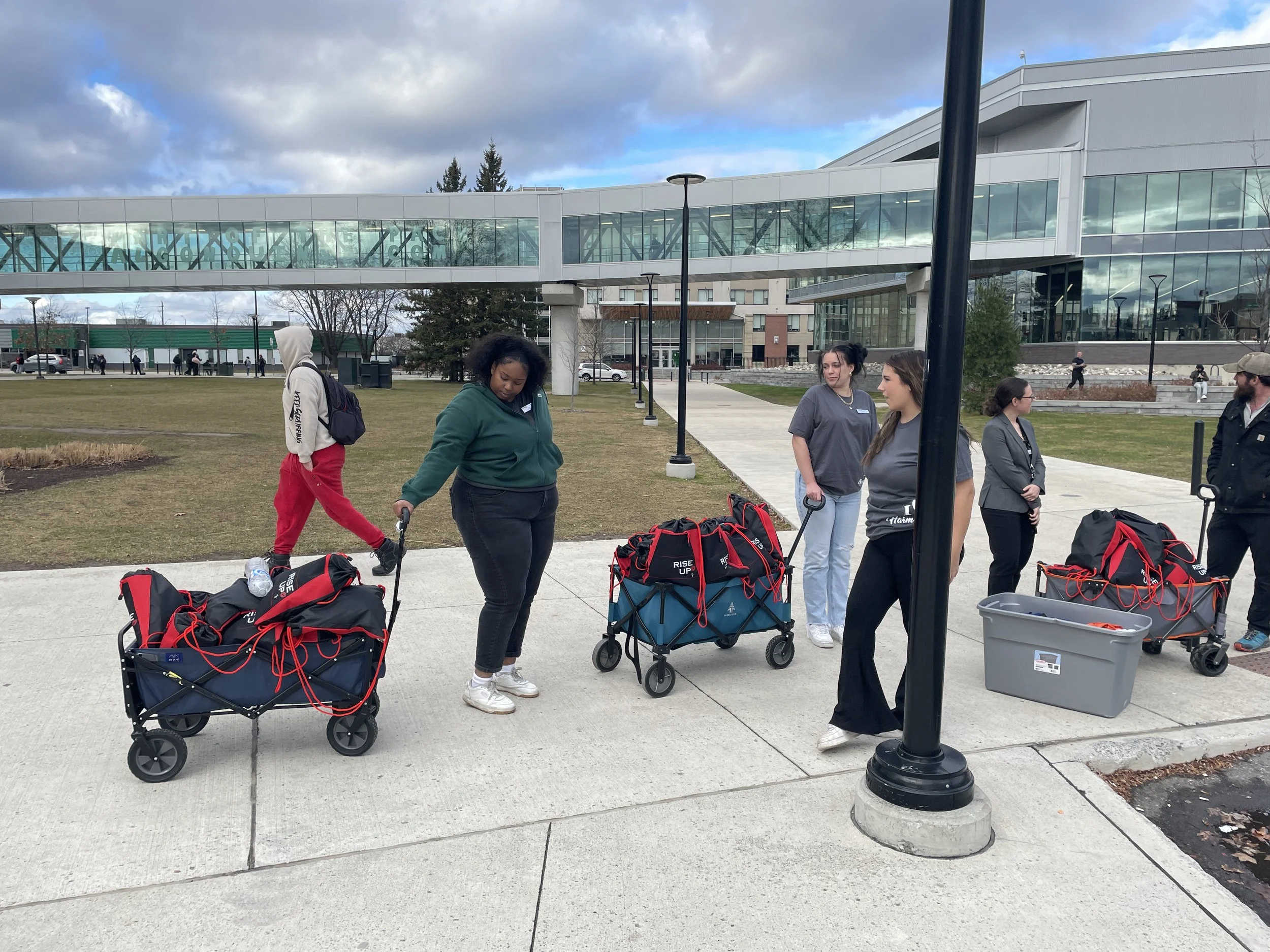 Group of people standing outside on a sidewalk with wagons filled with black and red duffel bags, near a modern glass building and grassy area.