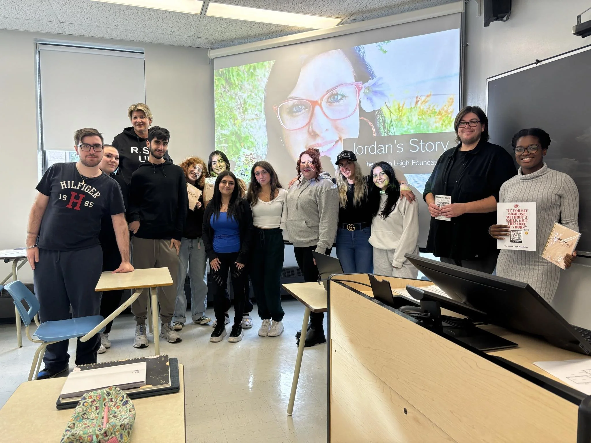 Group of students and a teacher standing in a classroom in front of a projection screen that reads 'Jordan's Story' with a photo of a smiling woman with red hair and glasses. The classroom has desks, a chalkboard, and various belongings on the desks.