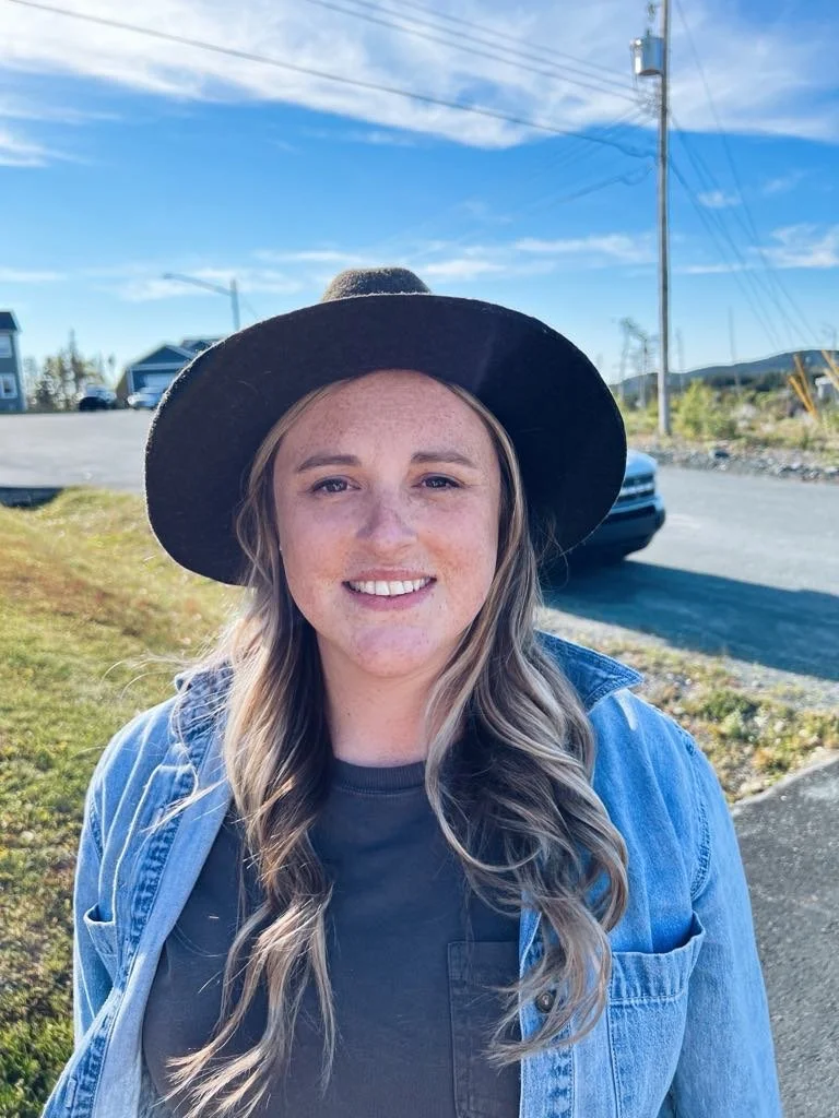 Young woman with long wavy blond hair, wearing a black wide-brimmed hat, a dark shirt, and a denim jacket, smiling outdoors on a sunny day with a clear blue sky, houses, and parked cars in the background.