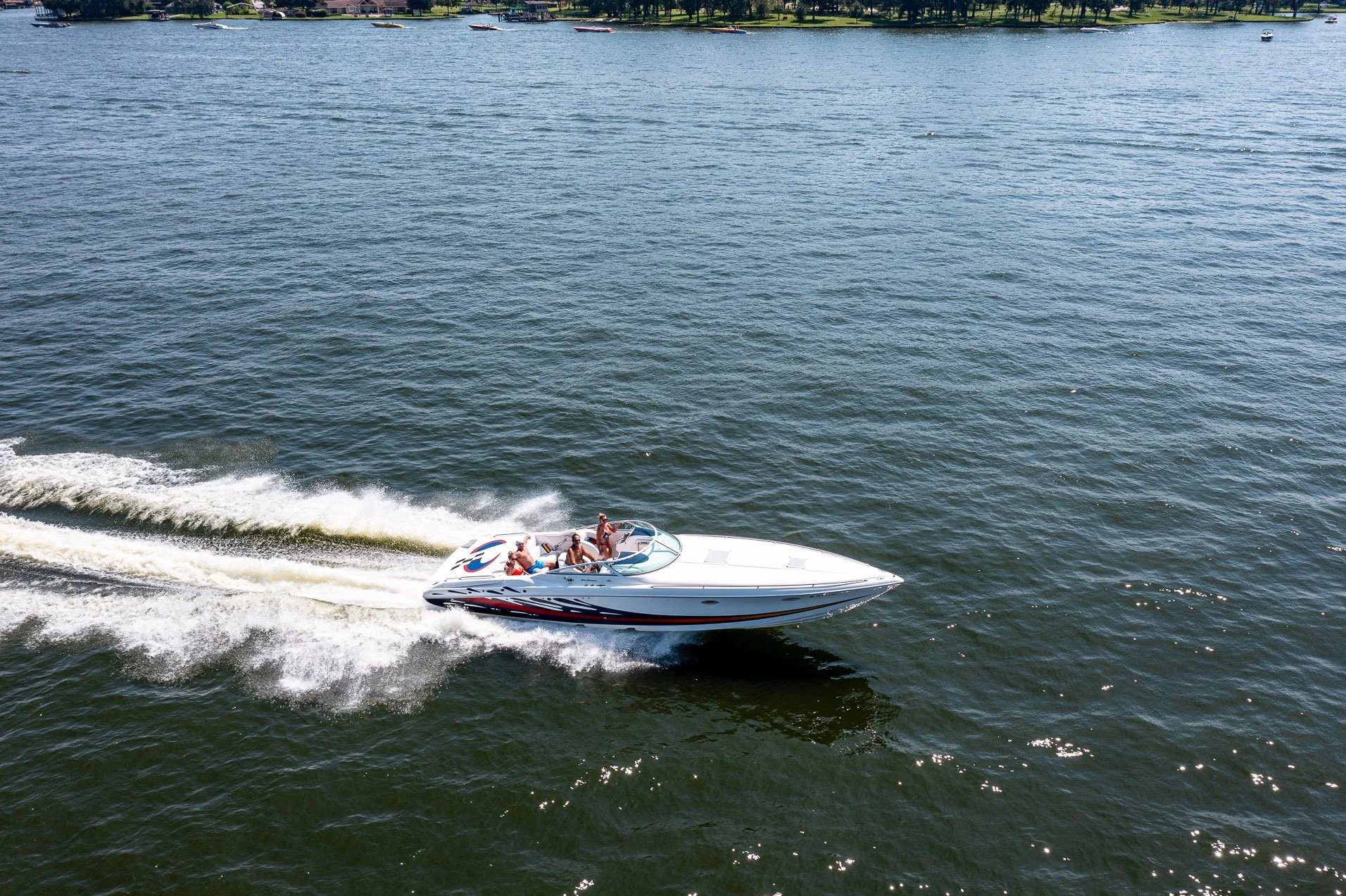 A group of four people enjoying a boat ride on a white speedboat in a large body of water, with trees and houses visible in the background.