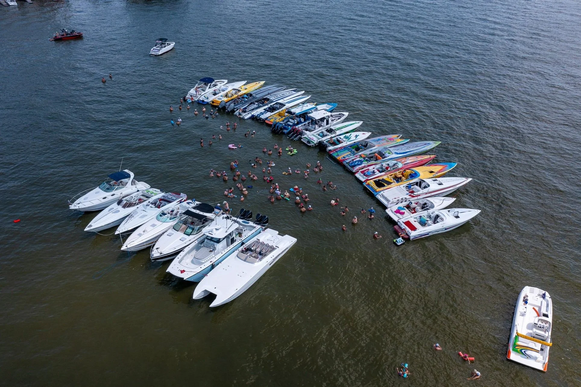 An aerial view of a marina with numerous boats docked in a semi-circle, and many people swimming and floating in the water nearby.