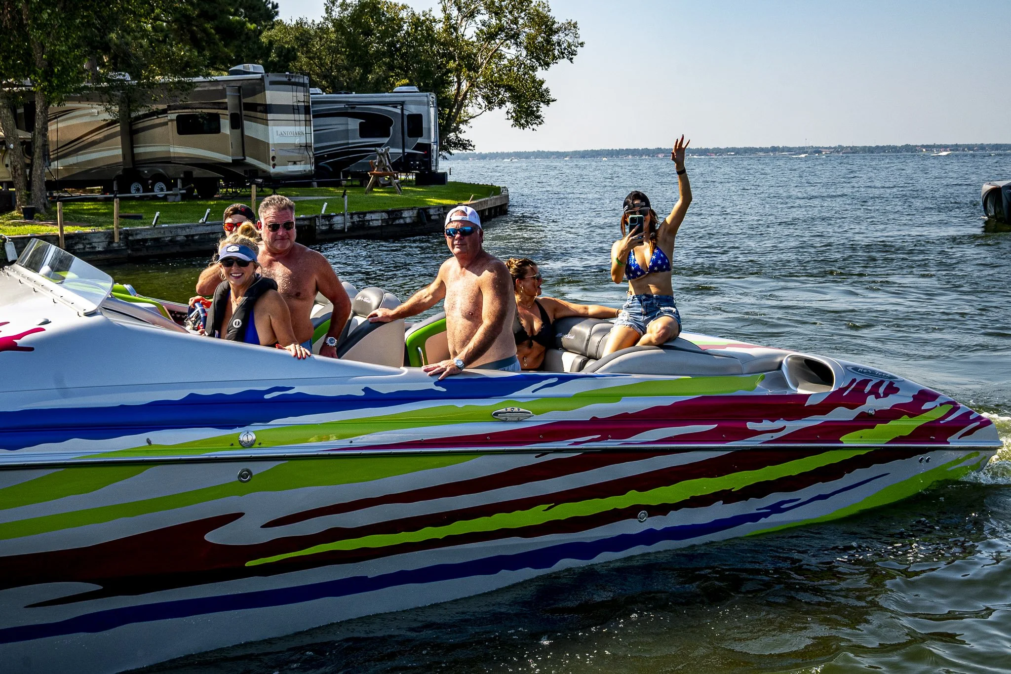 A group of six people on a colorful speedboat on a lake, with trees and a camper trailer in the background, enjoying a sunny day.