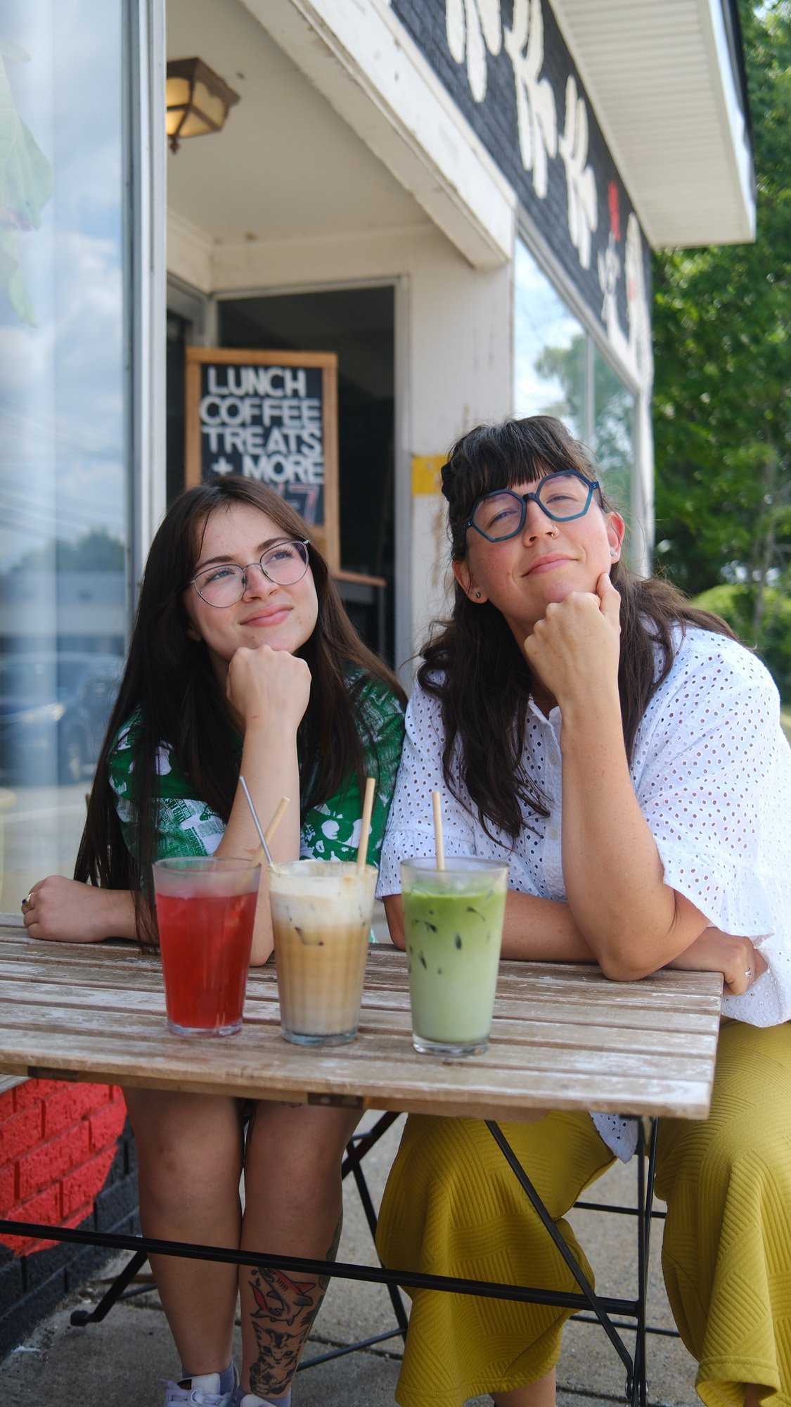 Two women wearing glasses pose at a café table with red, iced coffee, and green matcha drinks.