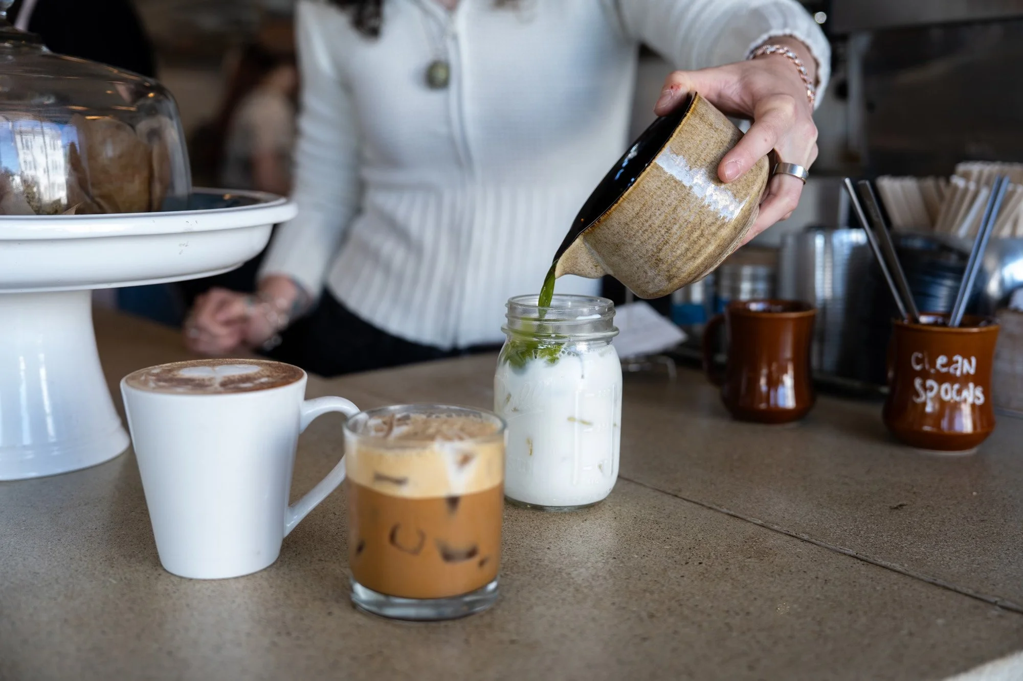 Barista pouring green matcha into a mason jar of milk beside coffee drinks at a cafe counter