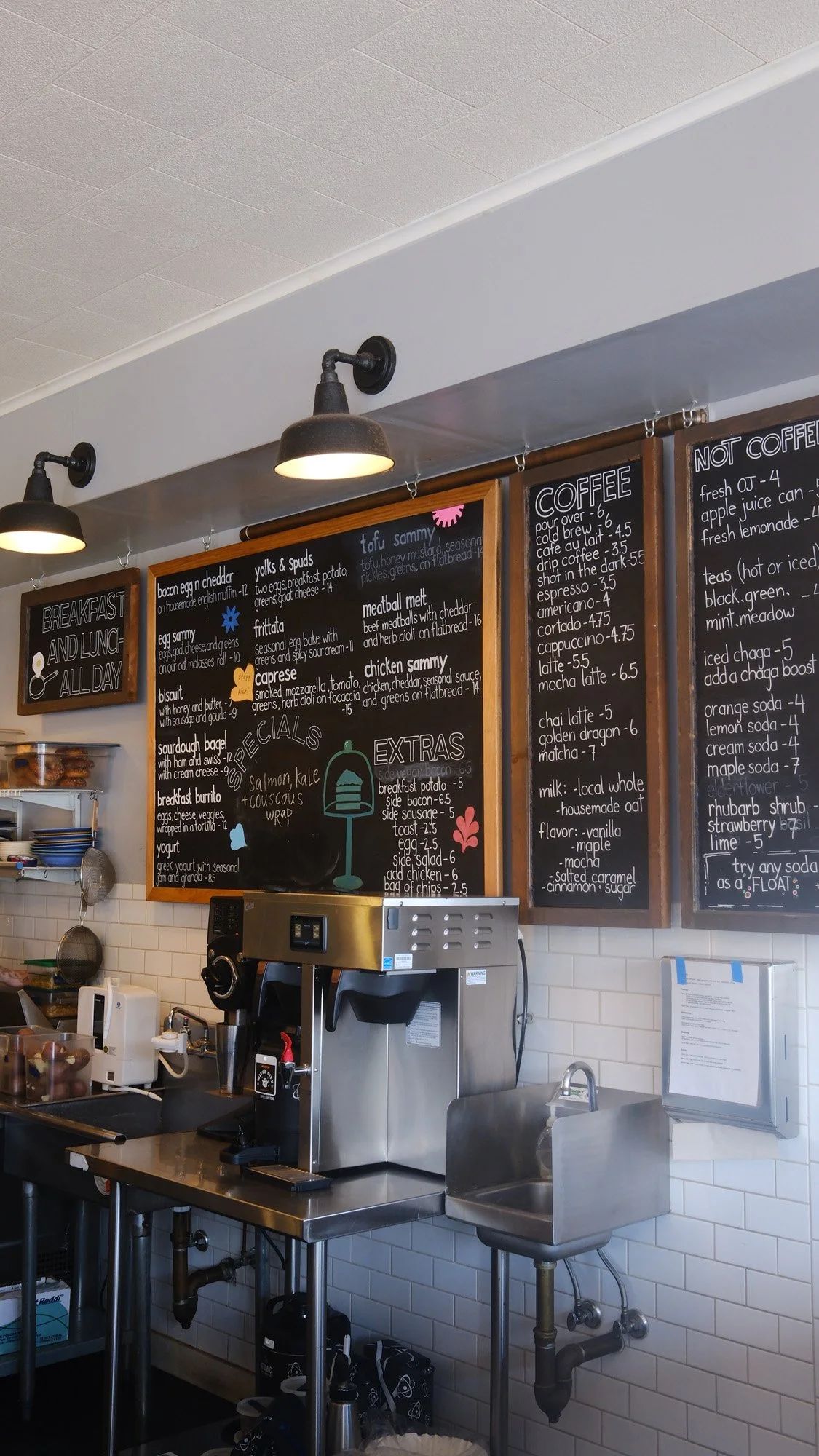Cafe counter with chalkboard menus, espresso machine, pendant lights, and stainless steel sink