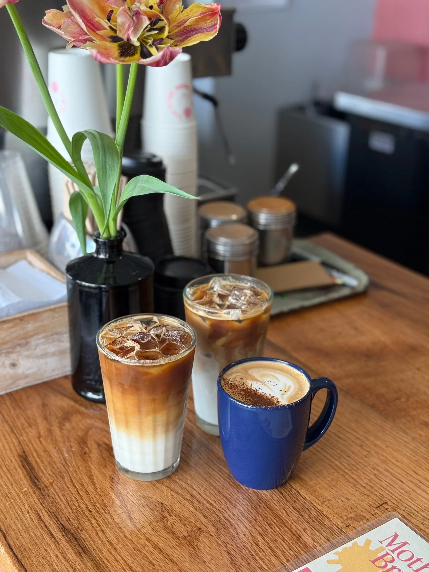 Three iced coffee drinks and a cappuccino on a cafe table beside a vase of flowers
