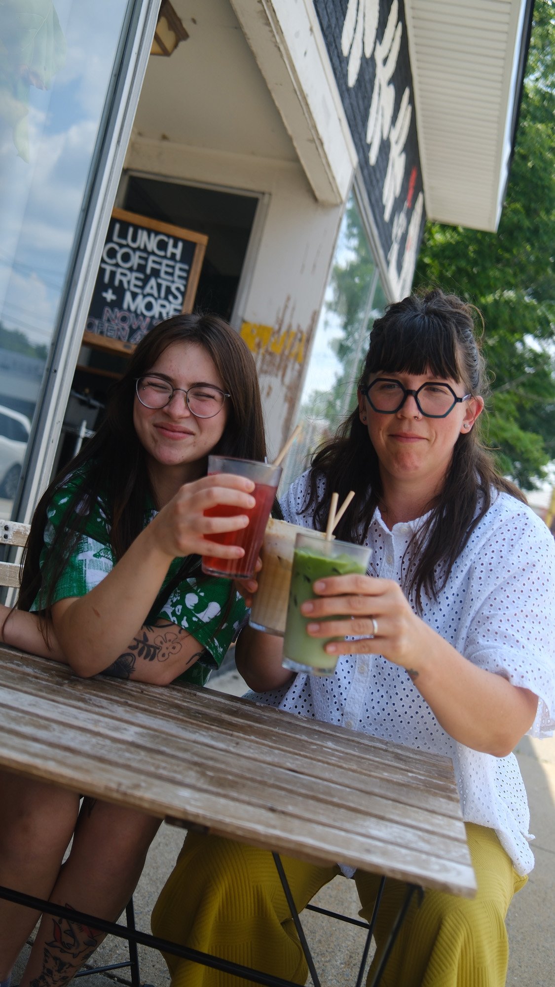 Two women wearing glasses enjoy colorful drinks at an outdoor café table.