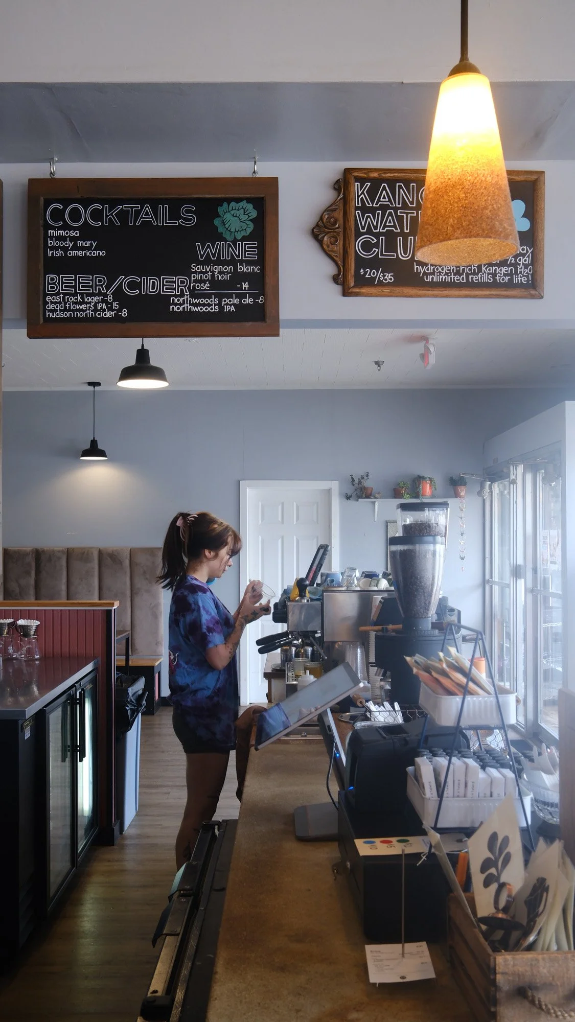 Barista preparing a drink behind a cafe counter with chalkboard menus, espresso machine, coffee grinder, and pendant lights.