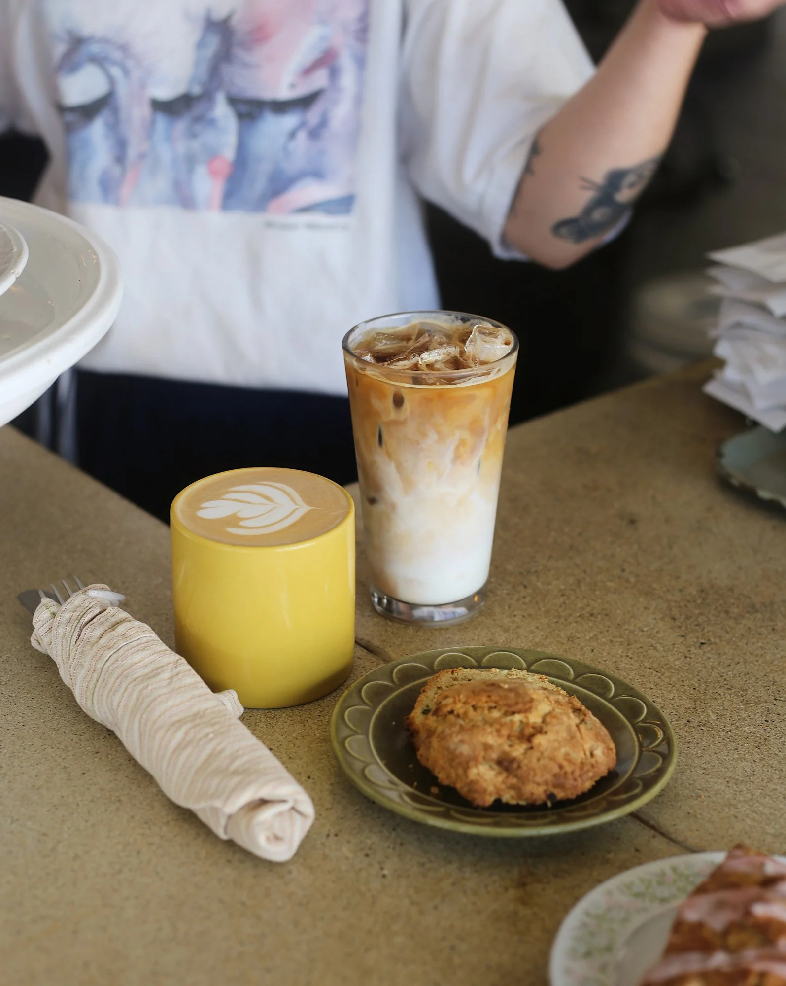 Café table with a yellow latte, iced coffee, pastry, and rolled napkin.