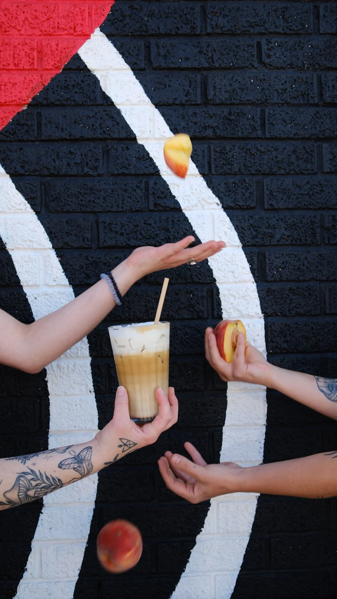 Hands holding a peach latte and sliced peaches in front of a black brick wall with white and red geometric paint