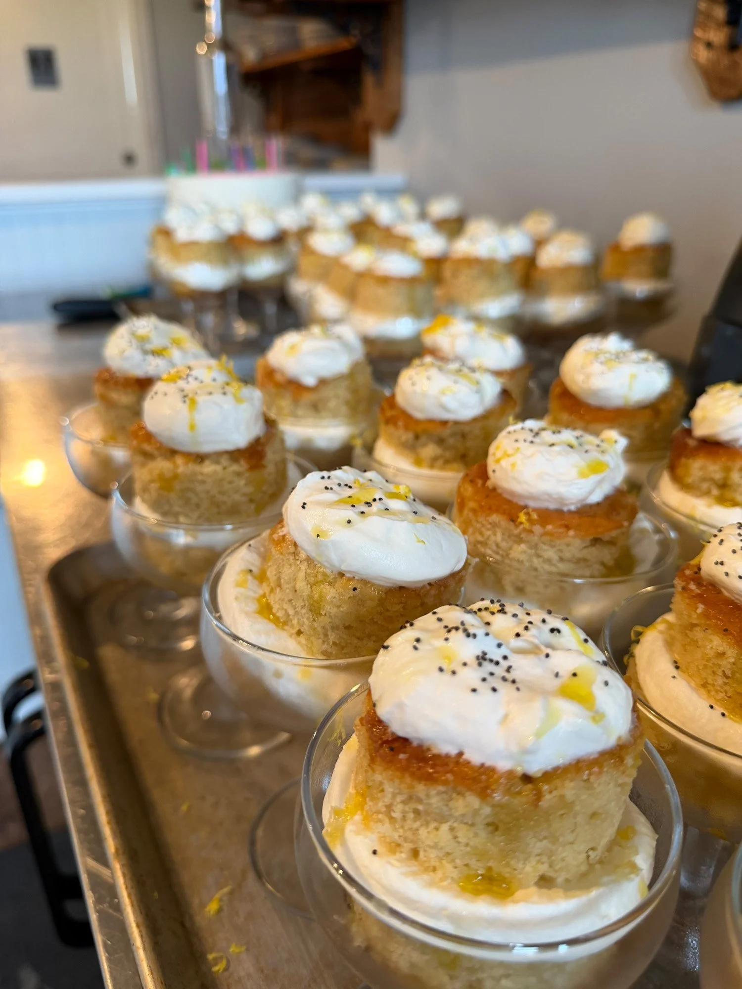 Mini lemon cakes topped with whipped cream and poppy seeds, served in clear glass cups on a tray