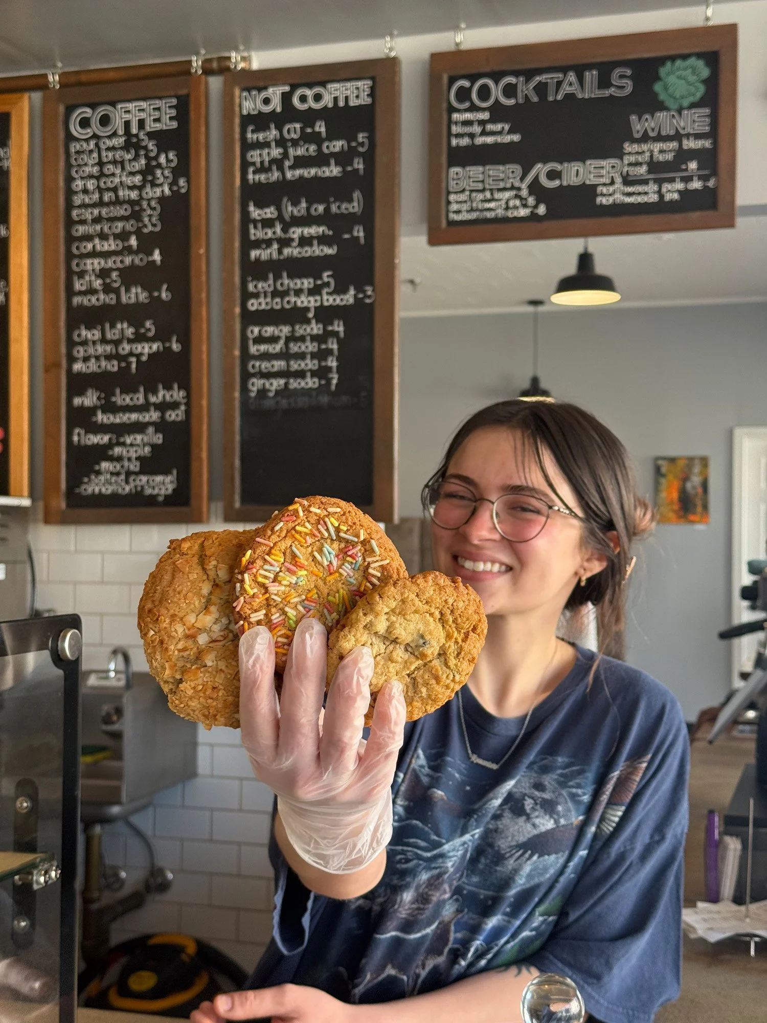Smiling barista in glasses and gloves holding a stack of oversized sprinkle-covered cookies in a cafe.