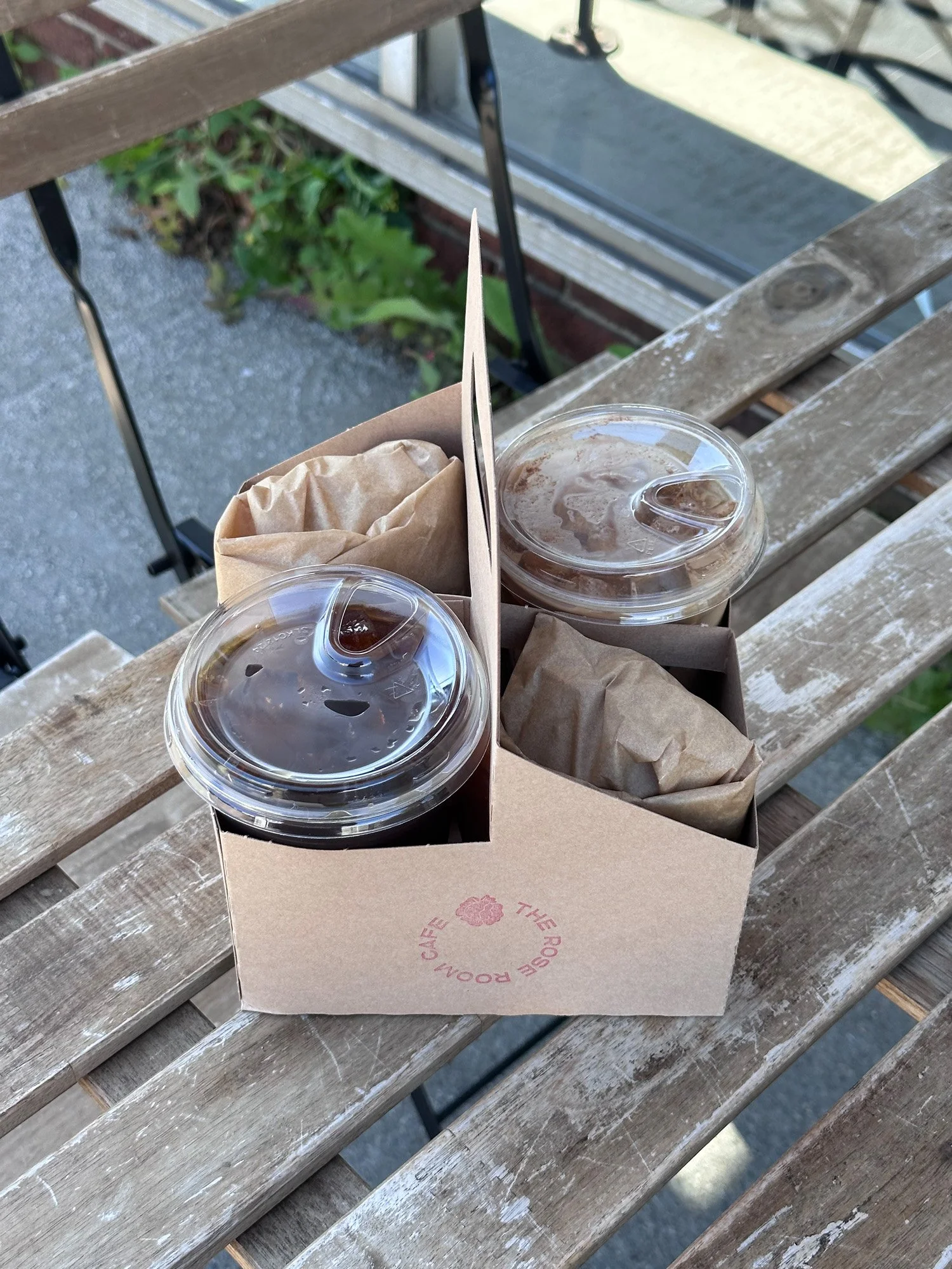 Two iced coffee drinks and wrapped pastries in a cardboard cafe carrier on a weathered outdoor picnic table.
