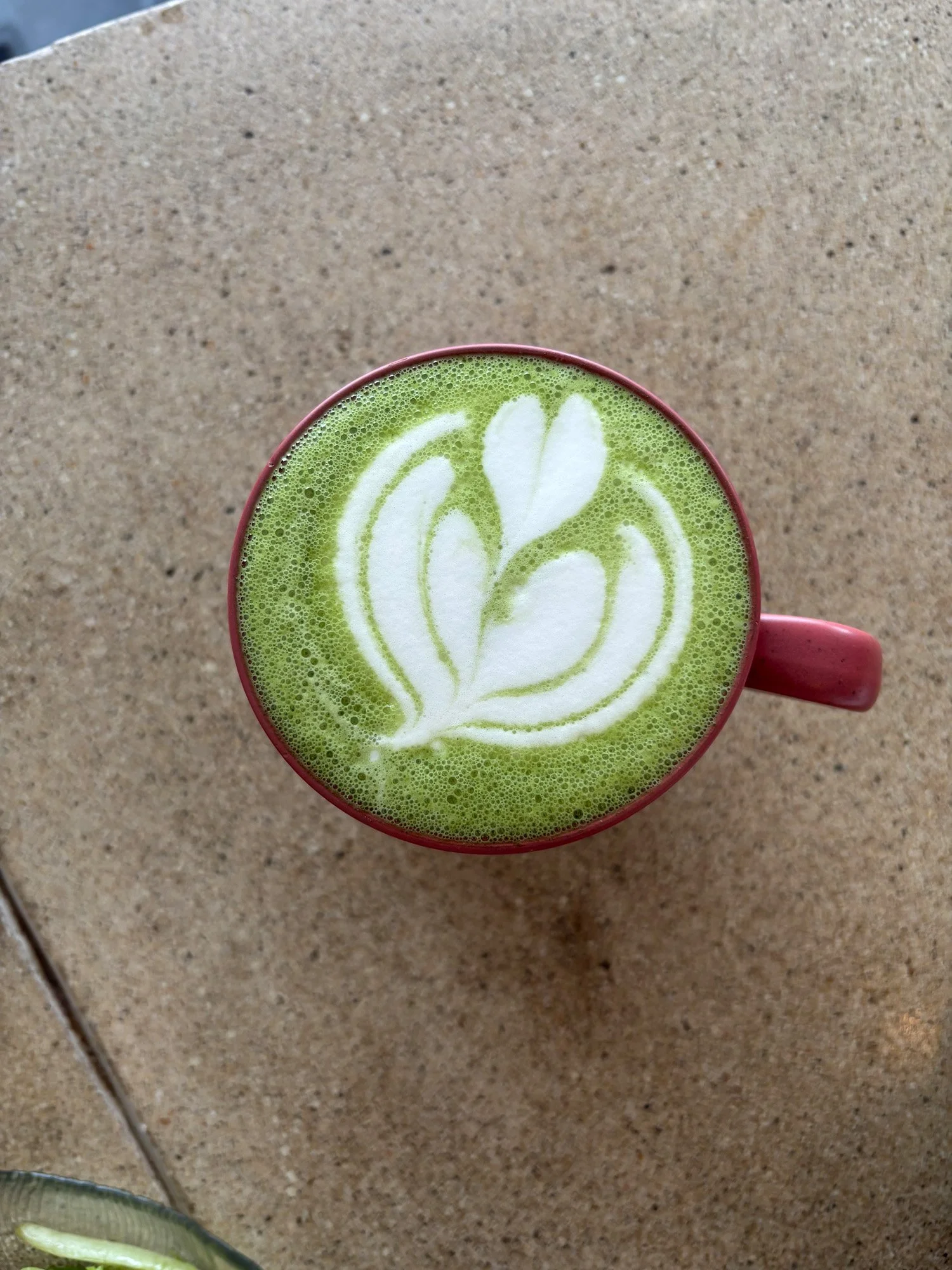 Top-down view of a red mug filled with green matcha latte and white leaf latte art on a stone tabletop