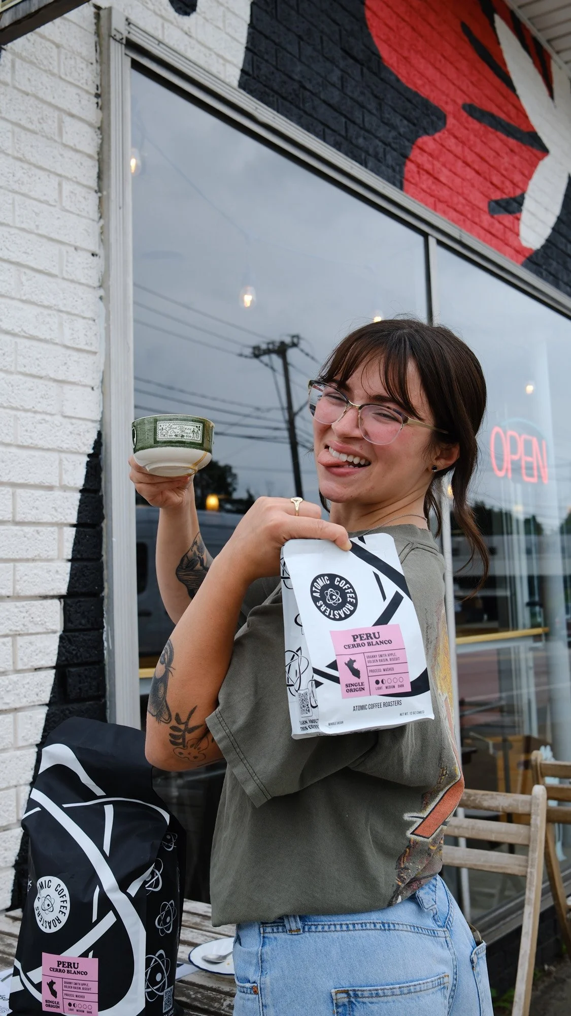 Smiling coffee shop employee holding a cup and Peru Cerro Blanco coffee bag outside a café with an open sign