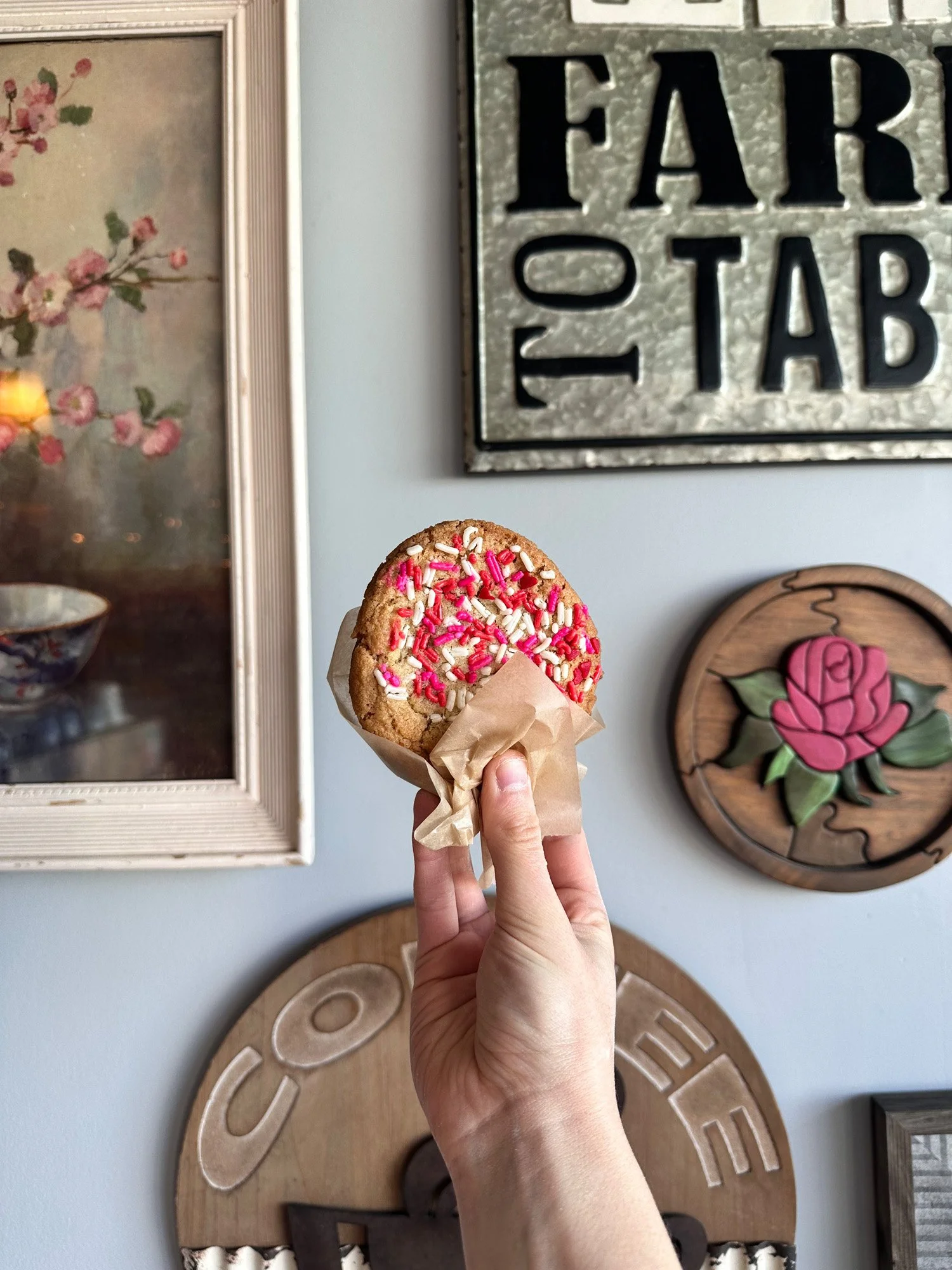 Hand holding a round sugar cookie covered in pink and white sprinkles against a wall decorated with framed floral art and vintage signs.