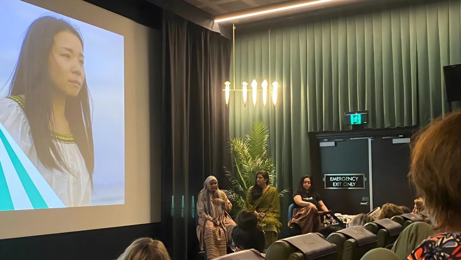 Panel discussion with Shuchi Kothari, Shreya Gejji and Mia Maramara, seated in front of a screen showing a still from 'Kainga'; audience seated in a theater with green curtains and palm plant.