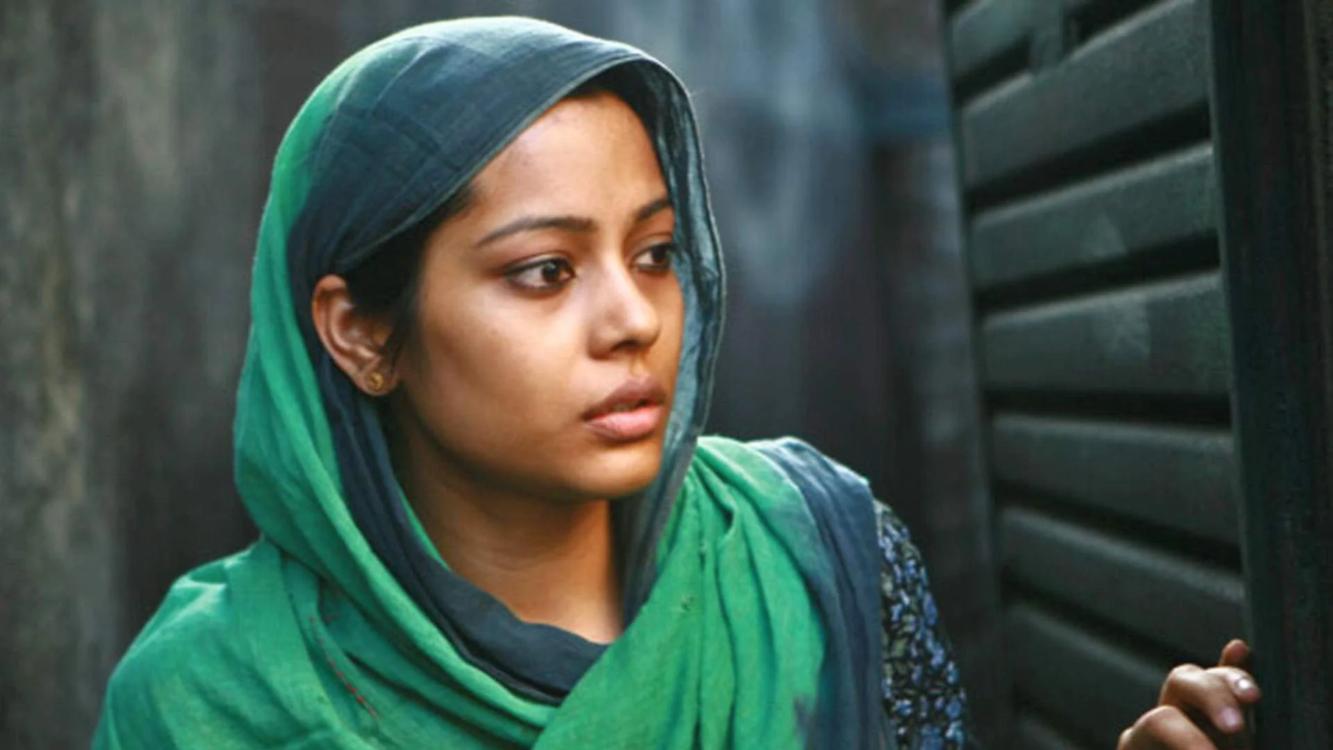A woman with a green headscarf and matching green shawl looks to the side with a serious expression. She is positioned next to a dark wall with horizontal vents.
