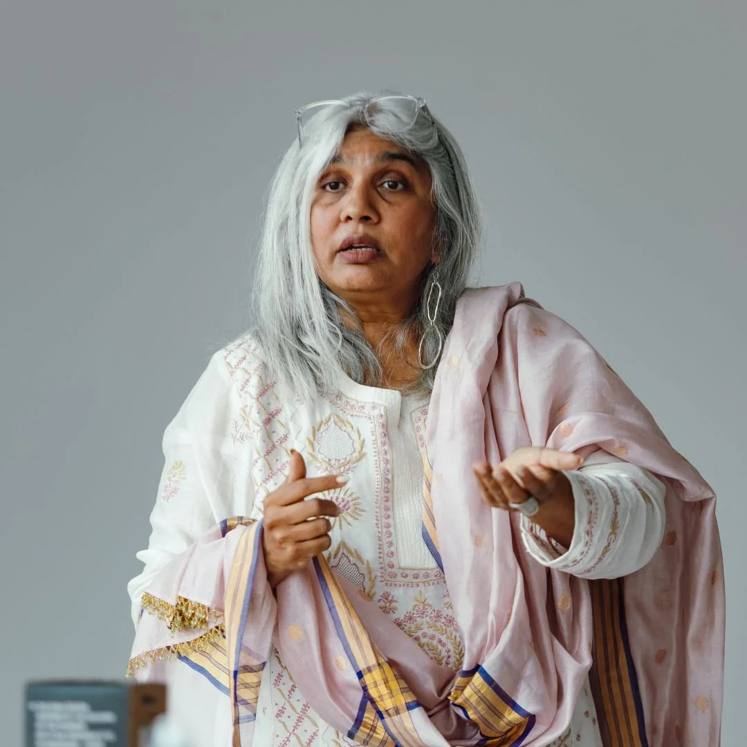 Shuchi Kothari wears a white sari with gold embroidery and a pink shawl, a light gray wall behind her, and gestures with her right hand.