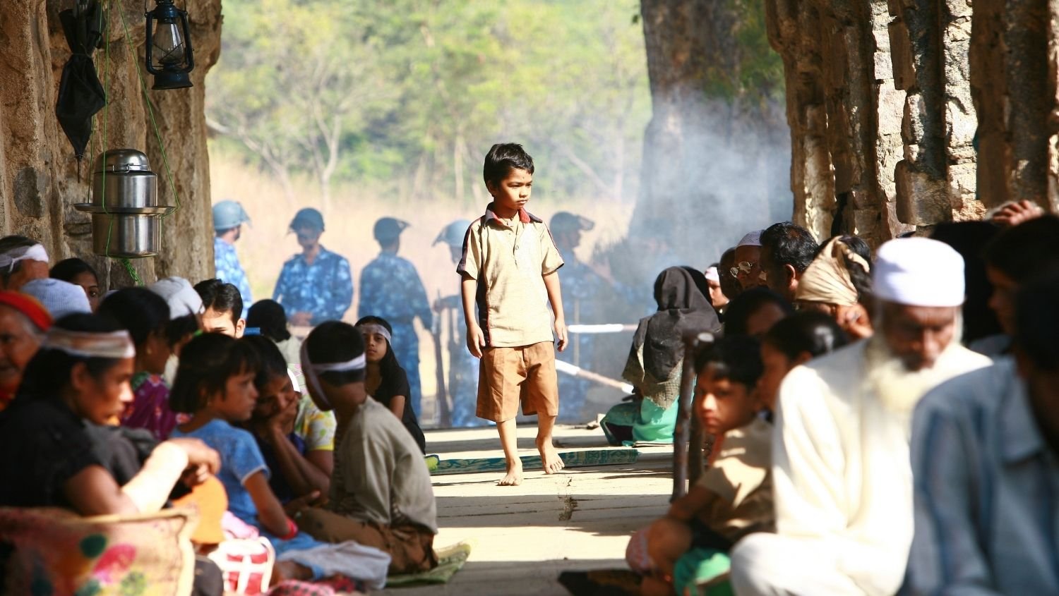 A boy walks barefoot in the center of a group of seated people inside a stone structure, with soldiers in blue camouflage uniforms and helmets visible outside in the background.