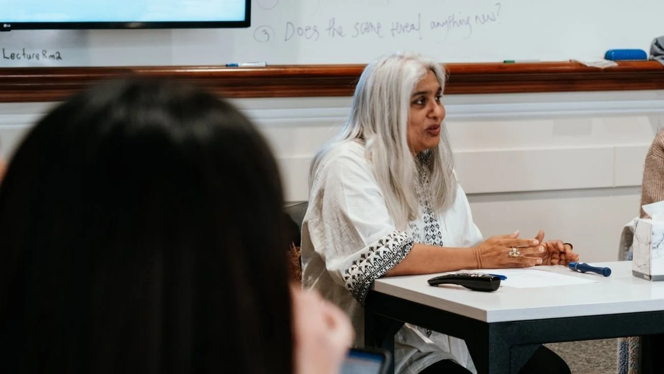 A woman with long gray hair speaking at a table in a classroom or meeting room, with a whiteboard behind her and other people partially visible in the foreground.