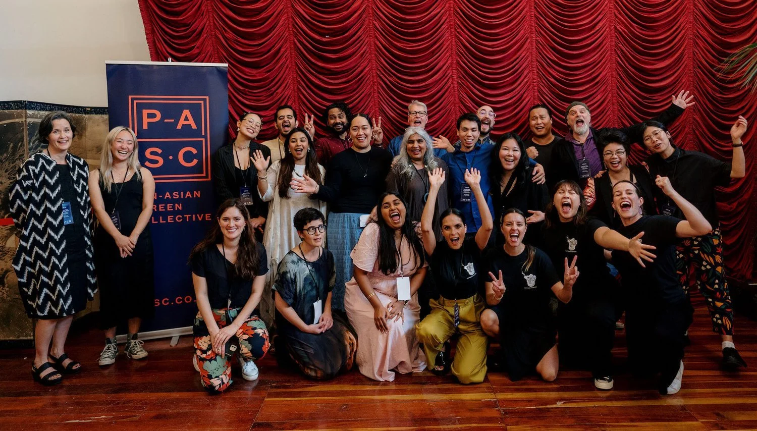 PASC members at an event, posing with a PASC banner, standing and kneeling in front of red curtain, smiling and celebrating.