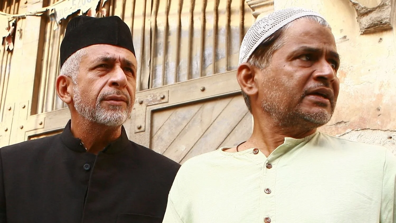 Two men with gray beards and middle eastern attire, one wearing a black cap and the other a white cap, standing outdoors near a beige building.