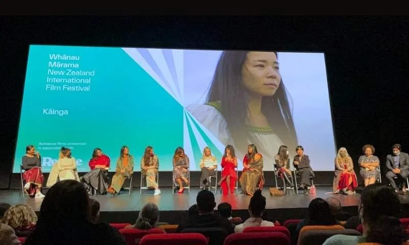 Panel of people sitting on stage during the Whānau Mārama New Zealand International Film Festival in Kãinga, with a large screen displaying a woman and festival information in the background.