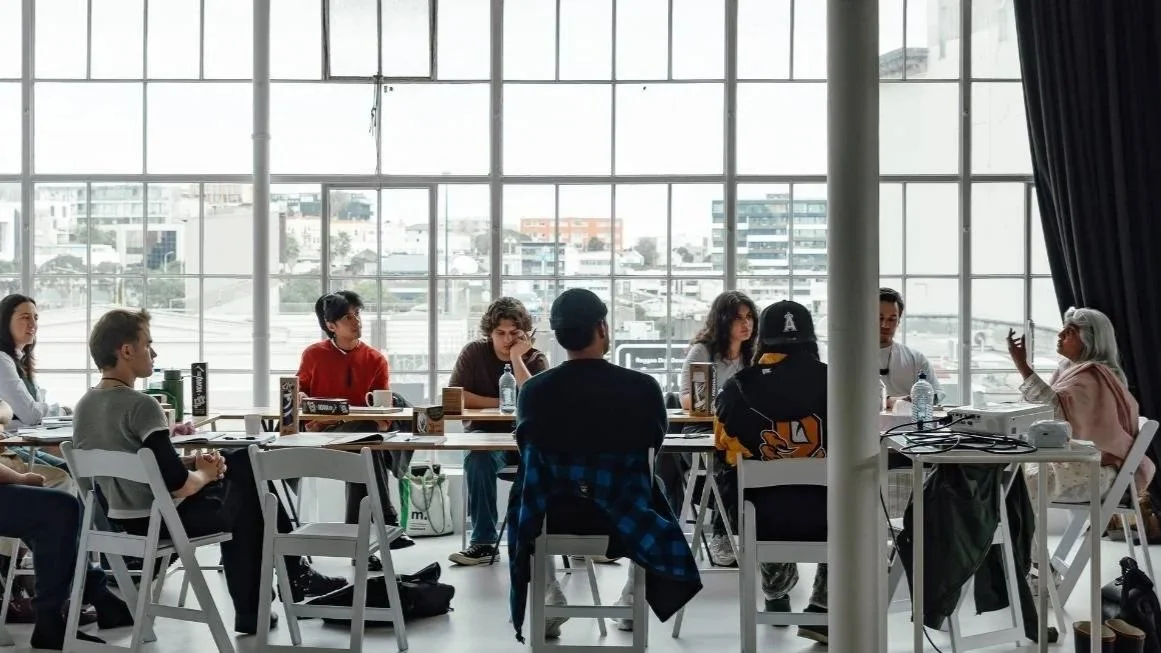 A group of lab participants sitting around a table in a modern conference room with large windows overlooking a cityscape. Shuchi Kothari presents to the group on the right of the image.