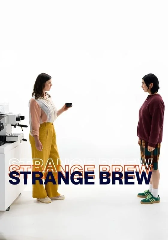 Poster for the web series 'Strange Brew'. Two women face each other in a white space with a coffee machine. One holds a cup of coffee. They both look awkward.