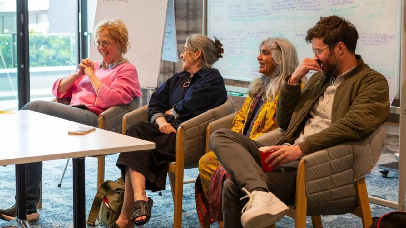 Participants in Writer’s Room: Seat at the Table - three women and one man - sit in chairs in a bright room with large windows. They laugh and engage in discussion, with notes and writing on a whiteboard behind them.