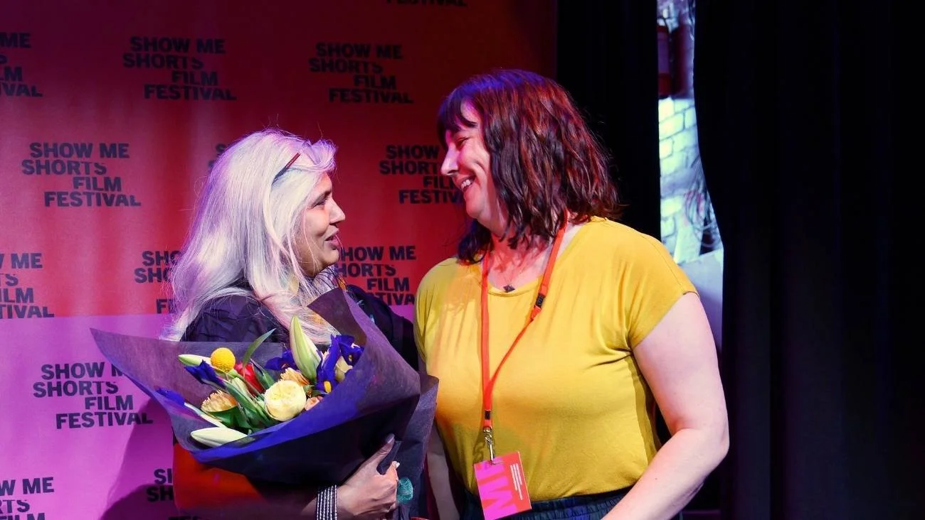 Shuchi Kothari holds a bouquet and smiles at Gina Dellabarca, in a yellow shirt and a red lanyard. Both are on the podium at the Show Me Shorts Film Festival.