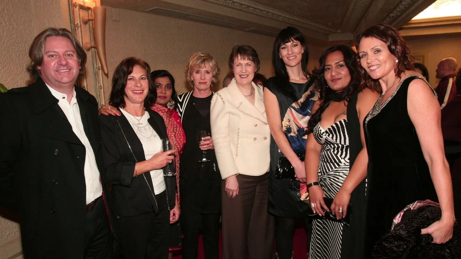 Group of eight women and one man at a social event, posing for a photo indoors, holding wine glasses, dressed in formal and semi-formal attire. Group includes former Prime Minister Helen Clark and Shuchi Kothari.