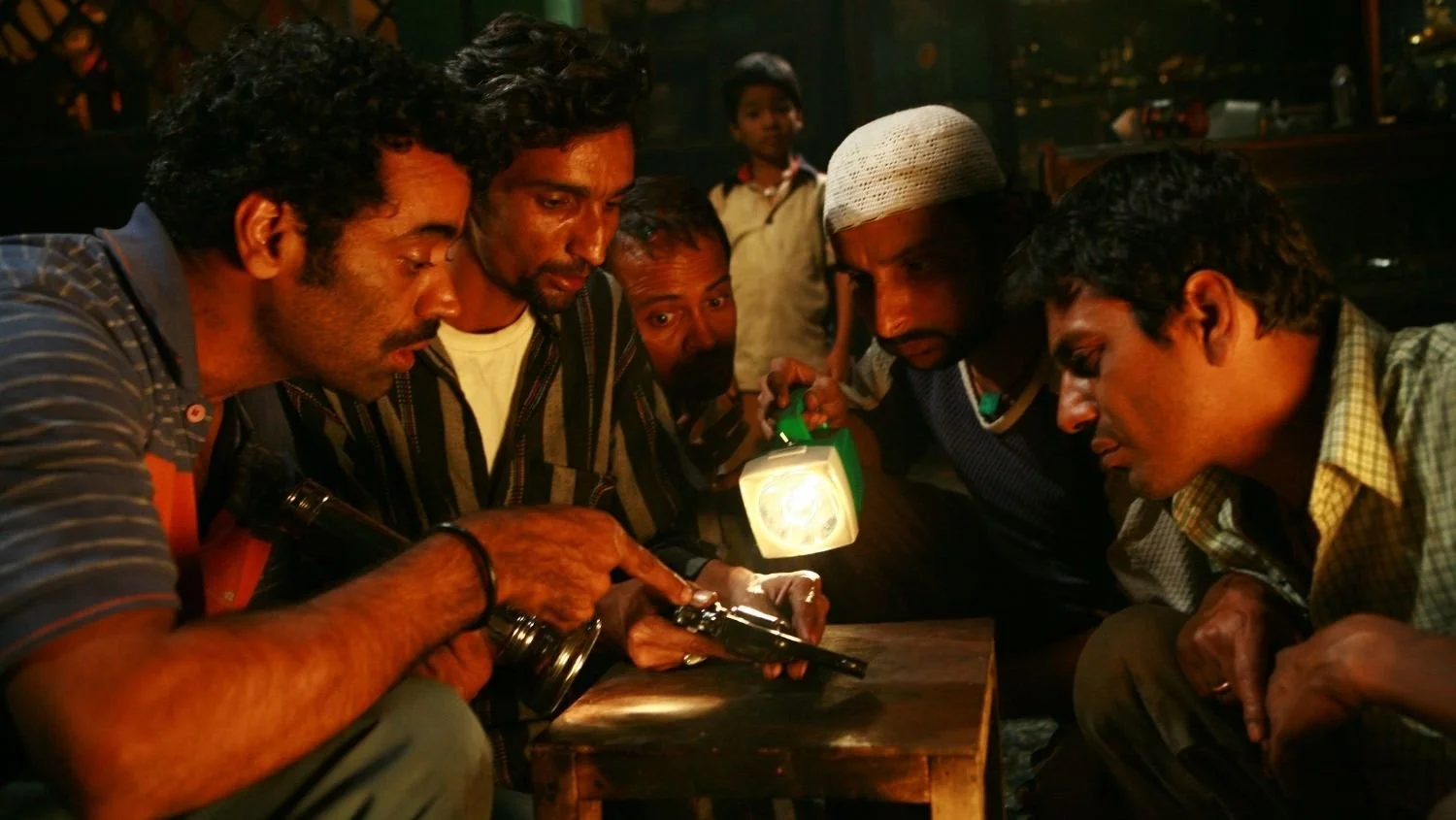 Group of six men closely gathered around a wooden table, examining a handgun under a flashlight at night.
