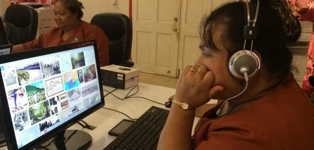A woman wearing headphones at a computer monitor displaying a collage of images, in a room with other people.
