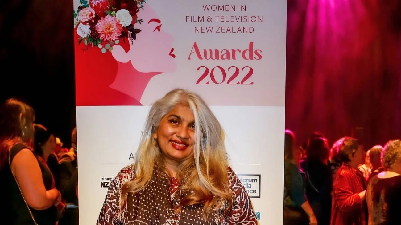 Shuchi Kothari smiling in front of a large pink and white poster at the Women in Film & Television New Zealand Awards 2022 event, with a crowd of women in the background.